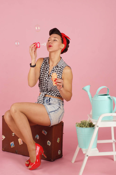 Young beautiful brunette woman sitting and blowing bubbles, pin-up style on pink background - Foto, Imagen