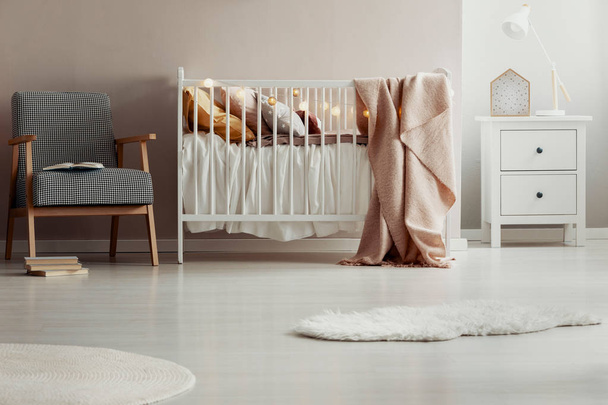 Stylish vintage armchair next to white wooden crib with pastel pink blanket, pillows and cotton ball lights - Foto, Imagen