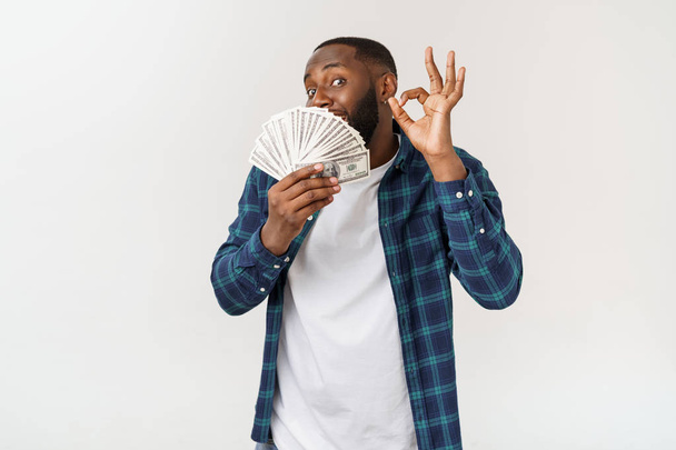 Portrait of young smiling dark skin african american man in white t shirt holding money and showing okay sign isolated on gray background - Fotó, kép