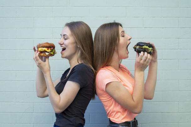 young beautiful girls-students stand near the blue wall, hold burgers and smile, they eat big cheeseburgers, opening their mouth wide - Valokuva, kuva