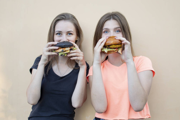 junge schöne Studentinnen stehen an der orangefarbenen Wand, halten Burger in der Hand und lächeln, sie freuen sich, wenn sie Fast Food essen - Foto, Bild