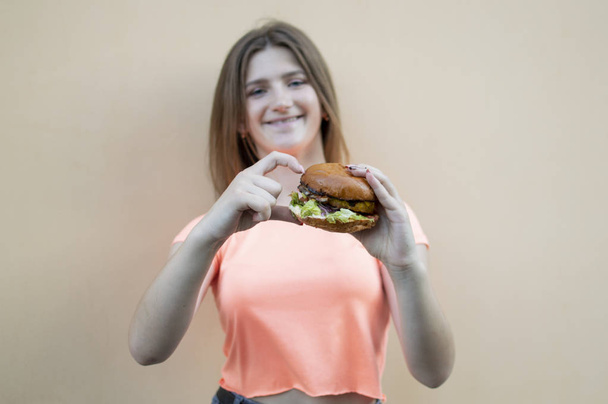 attractive teen girl stands against the background of an orange wall in an orange T-shirt and holds a big burger in her hand, she shows a heart with her fingers - Valokuva, kuva