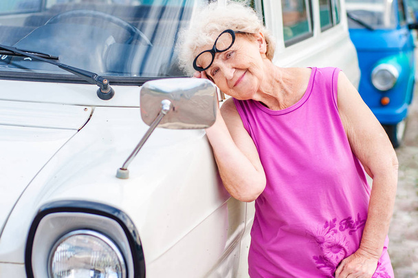 Модная привлекательная женщина в очках старше 60 лет смотрит на себя в зеркало ретро-машины. Летние каникулы и путешествия пожилых людей
 - Фото, изображение