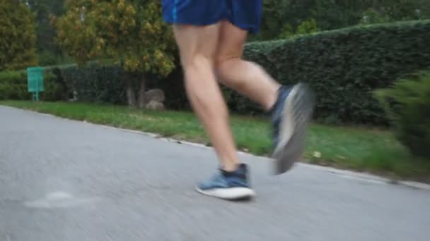 Athletic handsome man with wireless headphones jogging along sidewalk near forest at summer day. Young muscular sportsman running at nature. Concept of healthy active lifestyle. Crane shot Close up - Filmati, video