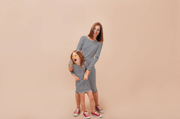6 yaşındaki kızı ile anne mutlu gerçek duyguları ile bej arka plan üzerinde poz aynı soyulmuş elbiseler giyiyor - Fotoğraf, Görsel