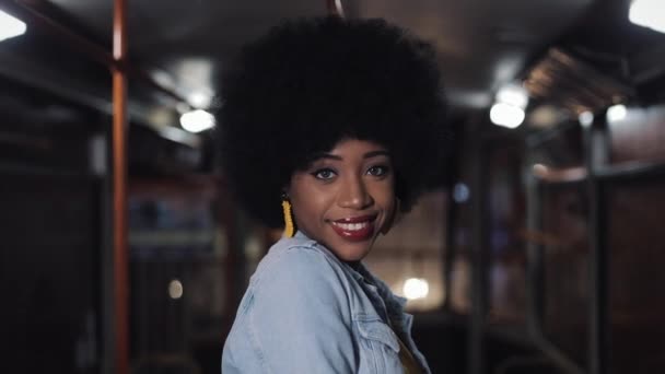 African american woman looking at the camera and smiling standing in the public transport, steadicam shot. Close-up. City lights background. - Filmati, video