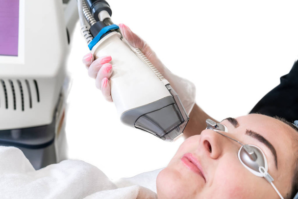 顔の若返りのために女性患者に対してレーザー治療を行う。白い背景とハンドピースを示しています。眼の保護を身に着けている患者. - 写真・画像