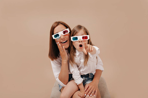 ziemlich charmantes kleines Mädchen mit Mama, die einen Film in 3D-Brille anschaut und einen Punkt vor die Kamera zeigt - Foto, Bild