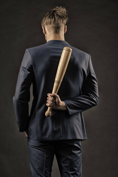 Latent aggression. Man with bat hides his aggression slow down and keep calm, rear view. Businessman or man in formal suit hides wooden bat behind back, dark background. Hidden danger concept - Foto, Imagem