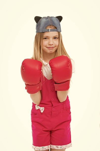 Lovely child wearing huge red boxing gloves. Girl in pink overalls isolated on white background. Kid wearing cute cap with animal ears backwards. Little fighter on training, self defence concept - Фото, изображение