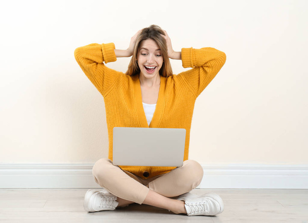 Surprised young woman with laptop sitting on floor near light wall indoors - Φωτογραφία, εικόνα