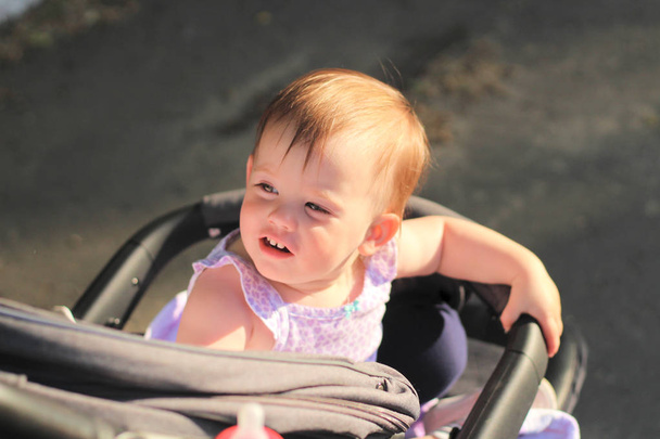 pieni, kaunis, hymyilevä, söpö punapää vauva hihaton paita vaunuissa ulkona putoaa kädet alas ja katselee taaksepäin
 - Valokuva, kuva