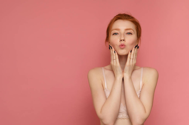 Redhead young woman wearing lace bra with a bun on head makes a kissy face on pink background - Фото, изображение