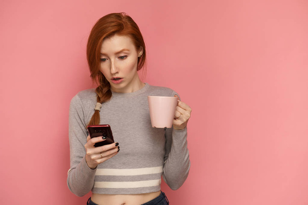 Red-haired surprised girl holds a cup in one hand looking at her smartphone in other hand - Photo, Image