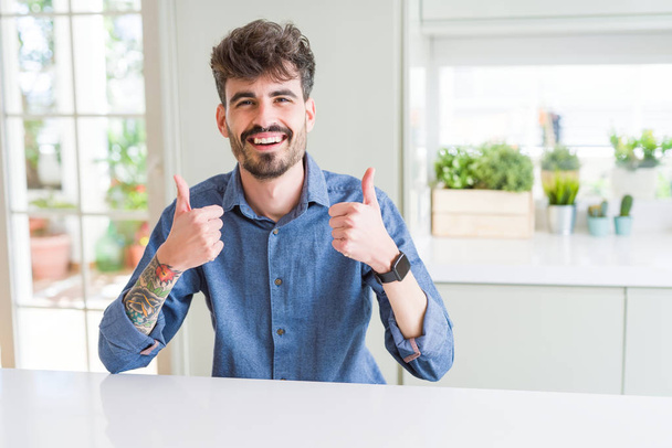 Young man wearing casual shirt sitting on white table success sign doing positive gesture with hand, thumbs up smiling and happy. Looking at the camera with cheerful expression, winner gesture. - Φωτογραφία, εικόνα