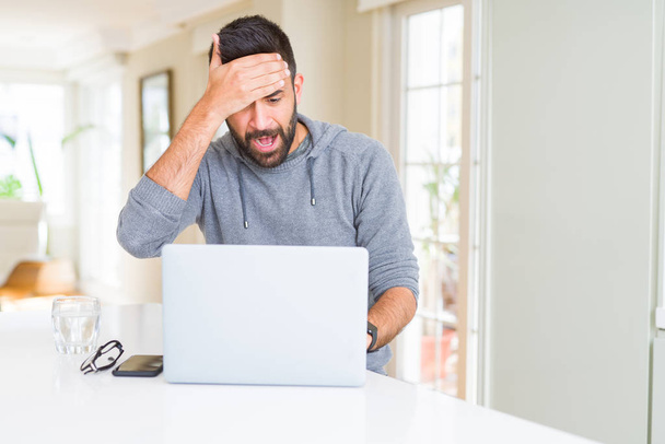 Hombre hispano guapo trabajando con computadora portátil estresado con la mano en la cabeza, conmocionado con vergüenza y cara sorpresa, enojado y frustrado. Miedo y malestar por error
. - Foto, Imagen