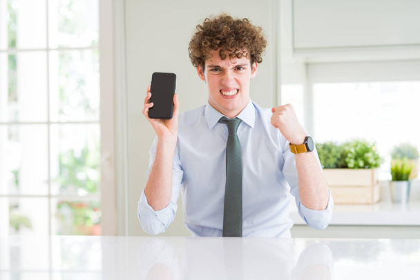 Νέοι επιχειρηματίες που δείχνουν smartphone οθόνη στο γραφείο ενοχλημένος και απογοητευμένοι φωνάζοντας με θυμό, τρελό και φωνάζοντας με ανυψμένο χέρι, έννοια του θυμού - Φωτογραφία, εικόνα