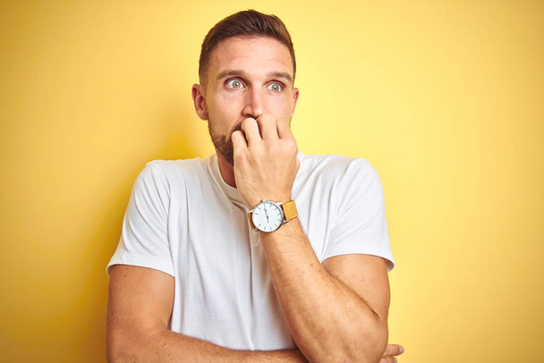 Jonge knappe man dragen casual wit t-shirt over gele geïsoleerde achtergrond op zoek gestrest en nerveus met handen op de mond bijten nagels. Angst probleem. - Foto, afbeelding