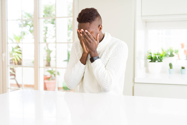 schöner afrikanisch-amerikanischer Mann auf weißem Tisch mit traurigem Gesichtsausdruck, der das Gesicht mit Händen bedeckt, während er weint. Depressionskonzept. - Foto, Bild