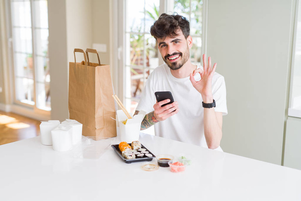 Молодий чоловік їсть азіатські суші з доставки додому і замовляє їжу, використовуючи додаток для смартфона, роблячи знак з пальцями, відмінний символ
 - Фото, зображення