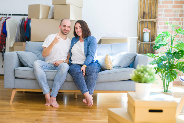 Молодая пара, сидящая на диване вокруг картонных коробок переезжая в новый дом показывая и показывая пальцем номер один улыбаясь уверенно и счастливо
. - Фото, изображение