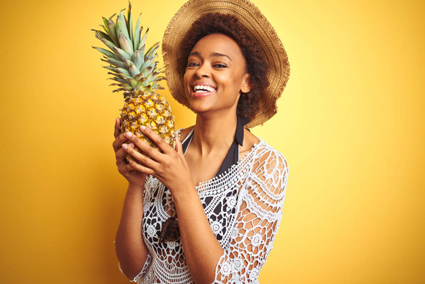 Jovem afro-americana com cabelo afro segurando pineaple sobre fundo amarelo isolado com um rosto feliz de pé e sorrindo com um sorriso confiante mostrando dentes
 - Foto, Imagem
