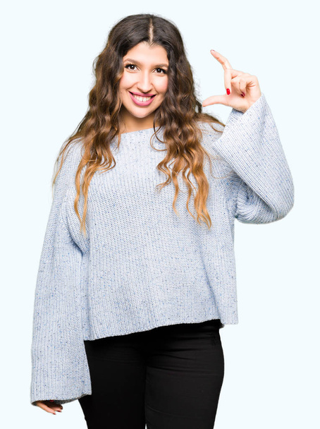 Молода красива жінка в зимовому светрі посміхається і впевнено жестикулює рукою, роблячи знак розміру пальцями, дивлячись і камеру. Концепція вимірювання
. - Фото, зображення