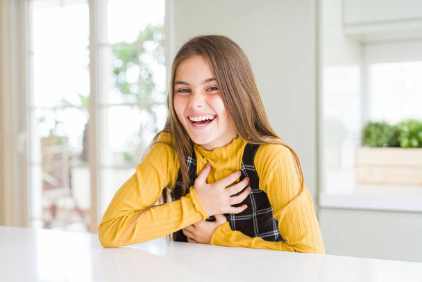 自宅でカジュアルな黄色のセーターを着て若い美しいブロンドの子供の女の子は、体に手で面白いクレイジージョークので、大声で笑って笑う. - 写真・画像