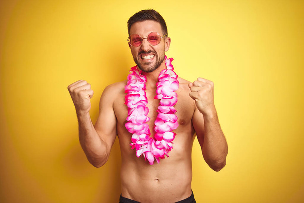 Young handsome shirtless man wearing sunglasses and pink hawaiian lei over yellow background very happy and excited doing winner gesture with arms raised, smiling and screaming for success. Celebration concept. - Photo, Image