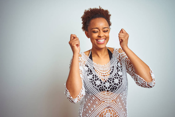 Fiatal afro-amerikai nő afro hajú visel bikini több mint fehér elszigetelt háttér izgatott a sikerhez a fegyverek emelt és a szemek zárva ünneplő győzelmét mosolyogva. Győztes koncepció. - Fotó, kép