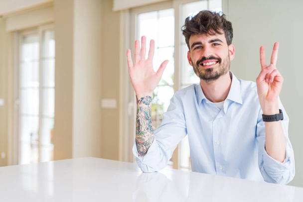 Jovem homem de negócios sentado na mesa branca mostrando e apontando para cima com os dedos número sete, sorrindo confiante e feliz
. - Foto, Imagem