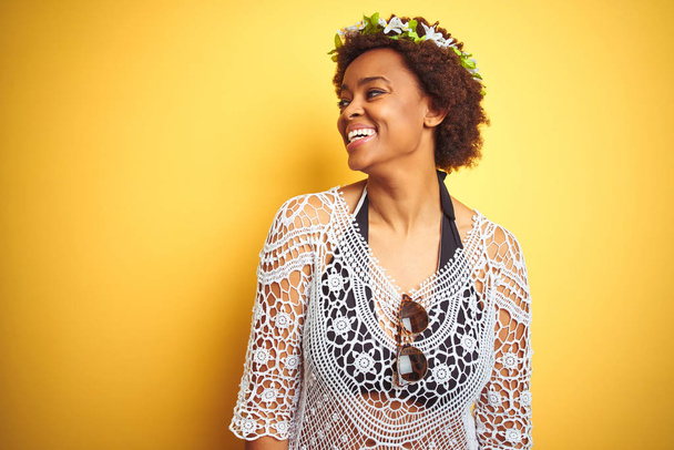 アフロヘアの若いアフリカ系アメリカ人女性は、黄色の孤立した背景の上に花の冠を身に着け、顔に笑顔で横を向き、自然な表情。自信を持って笑う. - 写真・画像