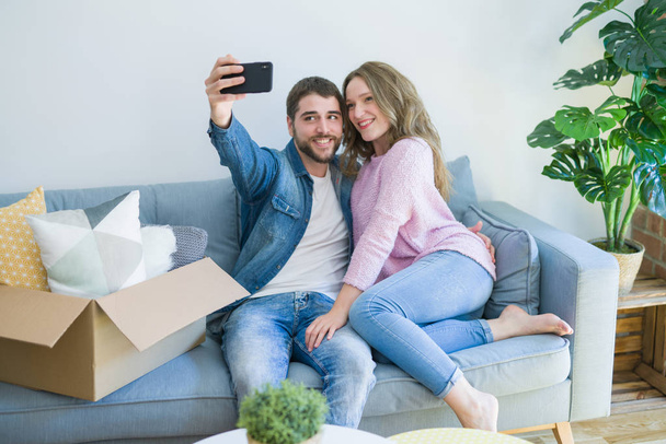 Молода красива пара фотографує селфі за допомогою смартфона, що сидить на дивані, посміхаючись дуже щаслива за переїзд в новий будинок
 - Фото, зображення