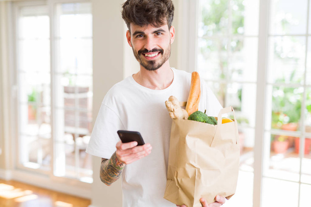 Mladý pohledný muž, který drží papírový pytel plný čerstvých potravin doma, zobrazuje a používá smartphone při nákupu produktů - Fotografie, Obrázek