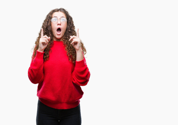 Beautiful brunette curly hair young girl wearing glasses and winter sweater over isolated background amazed and surprised looking up and pointing with fingers and raised arms. - Photo, Image
