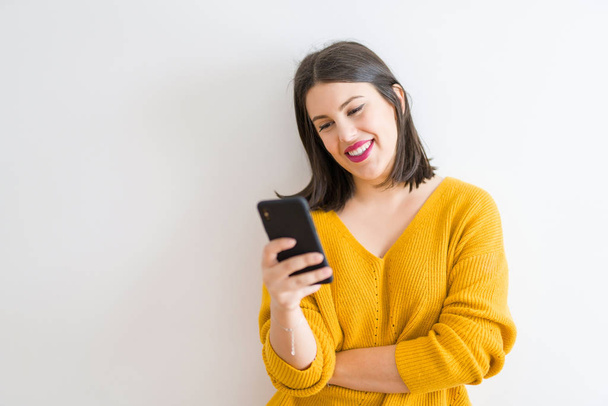 Kaunis brunette nuori nainen hymyilee luottavainen käyttämällä älypuhelinta yli eristetty valkoinen tausta yllään rento keltainen villapaita
 - Valokuva, kuva