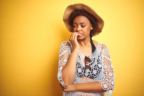 Молодая африканская американка с афроволосами в летней шляпе на белом изолированном фоне выглядит напряженной и нервной с руками во рту, грызущими ногти. Проблема тревоги
. - Фото, изображение