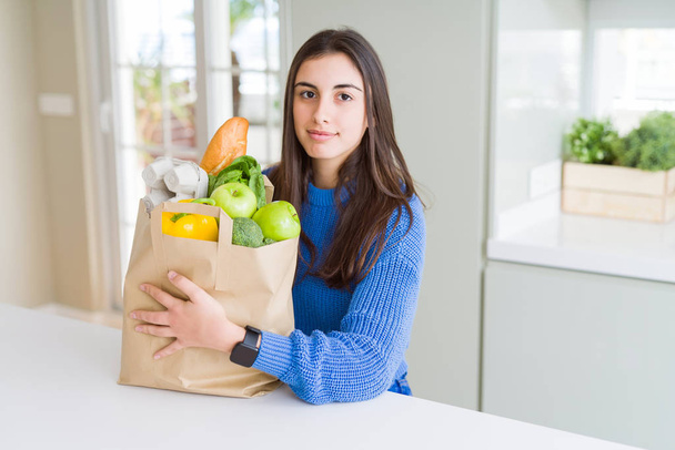 Красивая молодая женщина держит бумажный пакет, полный здоровых продуктов с уверенным выражением на умном лице, думая серьезно
 - Фото, изображение