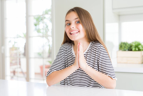 Красивая молодая девочка в полосах футболка молится с руками вместе просить прощения улыбаясь уверенно
. - Фото, изображение