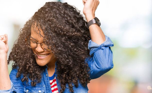 Young beautiful woman with curly hair wearing glasses very happy and excited doing winner gesture with arms raised, smiling and screaming for success. Celebration concept. - Photo, Image