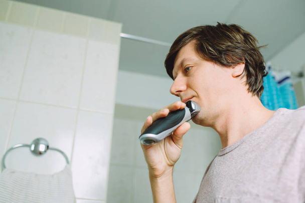 Fiatalember borotválkozás arcát usuNg egy elektromos borotvát a fürdőszobában - Fotó, kép