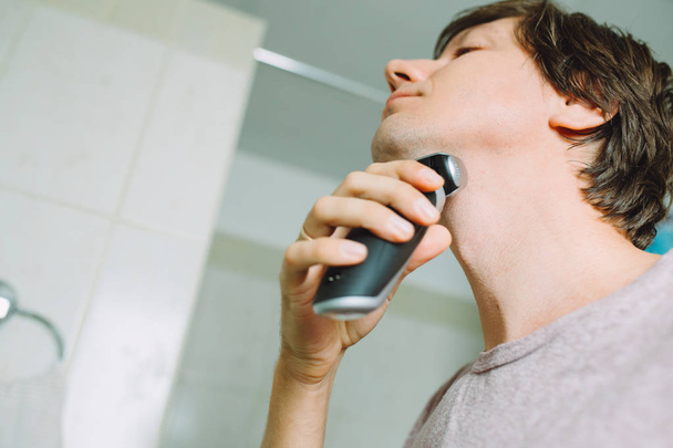 Νέος άντρας ξυρίζει το πρόσωπό του και ένα ηλεκτρικό ξυράφι στο μπάνιο - Φωτογραφία, εικόνα