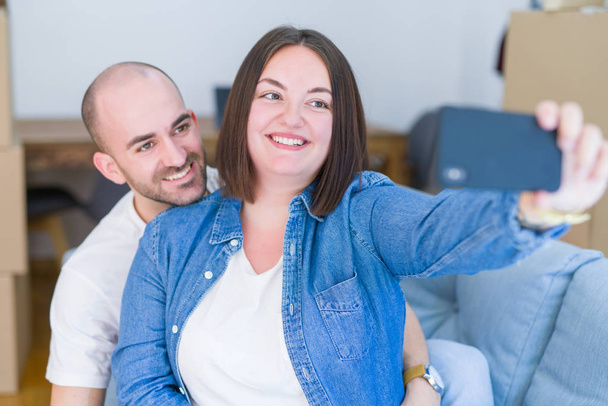Νεαρό ζευγάρι που κάθεται στον καναπέ στο νέο σπίτι παίρνοντας μια φωτογραφία selfie χρησιμοποιώντας smartphone χαμογελώντας χαρούμενος για τη μετακίνηση σε ένα νέο διαμέρισμα - Φωτογραφία, εικόνα