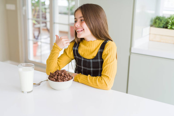 Beautiful young girl kid eating chocolate cereals and glass of milk for breakfast smiling with happy face looking and pointing to the side with thumb up. - Photo, Image