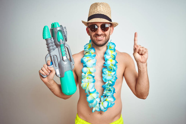 Mladý muž nosí letní čepici a havajské květiny, které drží vodní pistoli nad izolovaným pozadím, překvapené myšlenkou nebo otázkou ukazující prstem se spokojenou tváří, číslo jedna - Fotografie, Obrázek