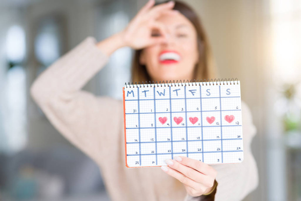Молода красива жінка тримає календар менструацій вдома з щасливим обличчям, посміхаючись роблячи знак ОК з рукою на око дивиться через пальці
 - Фото, зображення