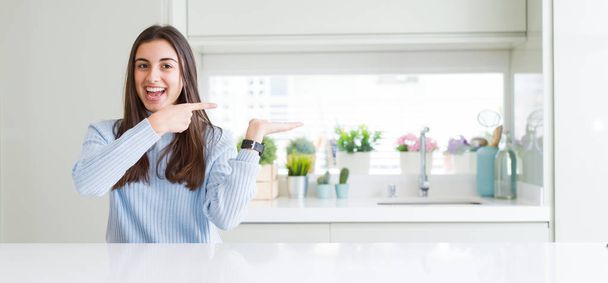 Wide angle picture of beautiful young woman sitting on white table at home amazed and smiling to the camera while presenting with hand and pointing with finger. - Фото, зображення