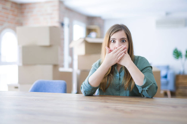 Young woman sitting on the table with cardboard boxes behind her moving to new home shocked covering mouth with hands for mistake. Secret concept. - Foto, imagen