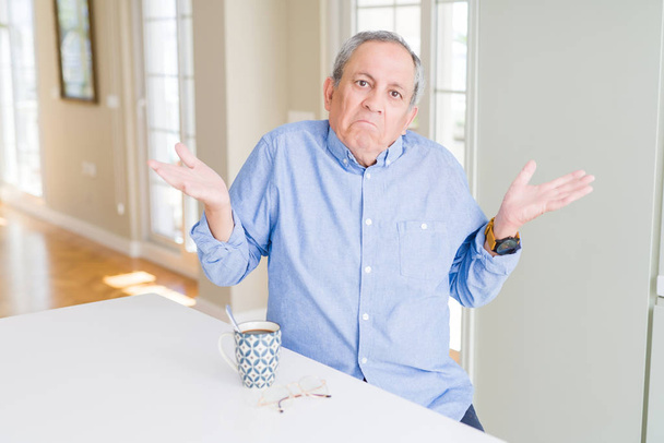 Yakışıklı yaşlı adam evde bir fincan kahve içme habersiz ve kolları ve elleri ile karışık ifade kaldırdı. Şüphe kavramı. - Fotoğraf, Görsel
