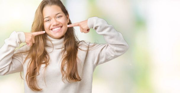 Young beautiful brunette woman wearing turtleneck sweater over isolated background smiling confident showing and pointing with fingers teeth and mouth. Health concept. - Fotó, kép